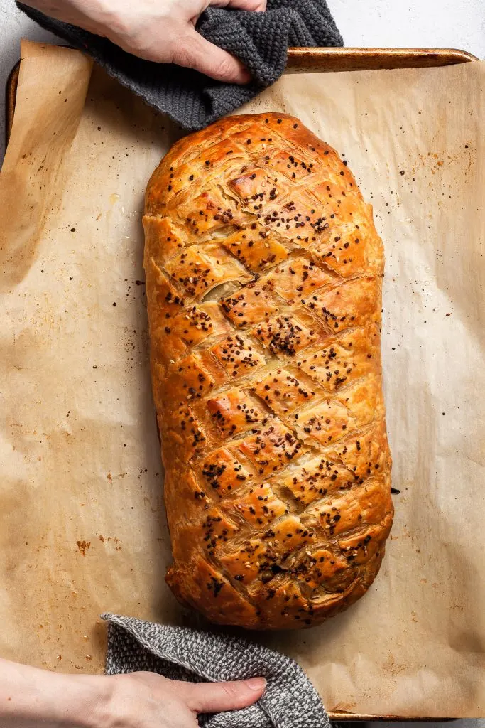 Vegan Wellington on a baking sheet hot from the oven being carried with grey knitted pot holders.