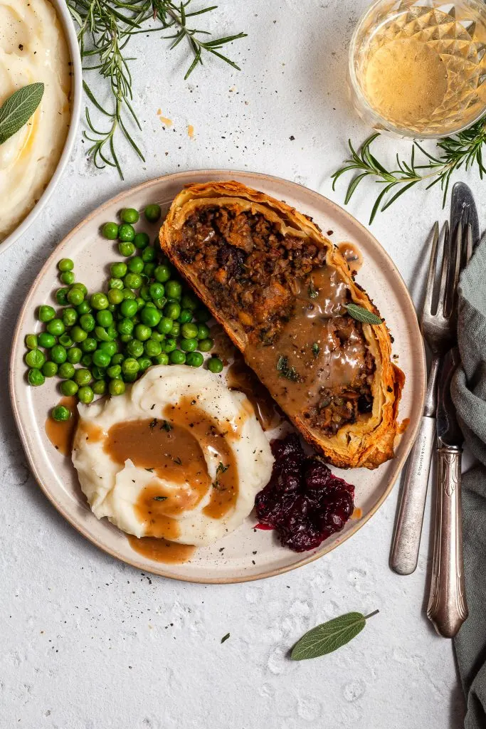A slice of vegan Wellington on a plate with mashed potatoes, green peas, cranberry sauce and gravy.