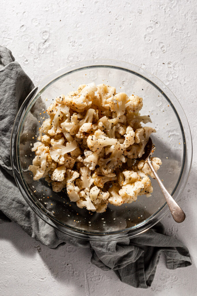 A clear glass bowl is filled with uncooked cauliflower florets which have spices and an oil like sheen covering them. 