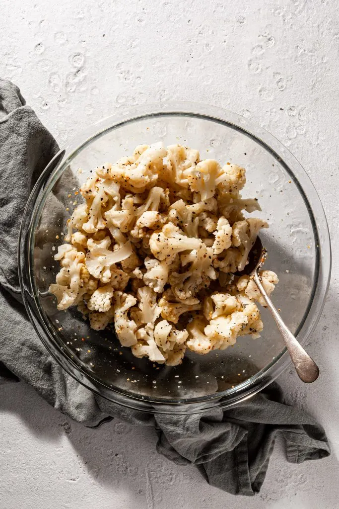 A clear glass bowl is filled with uncooked cauliflower florets which have spices and an oil like sheen covering them. 