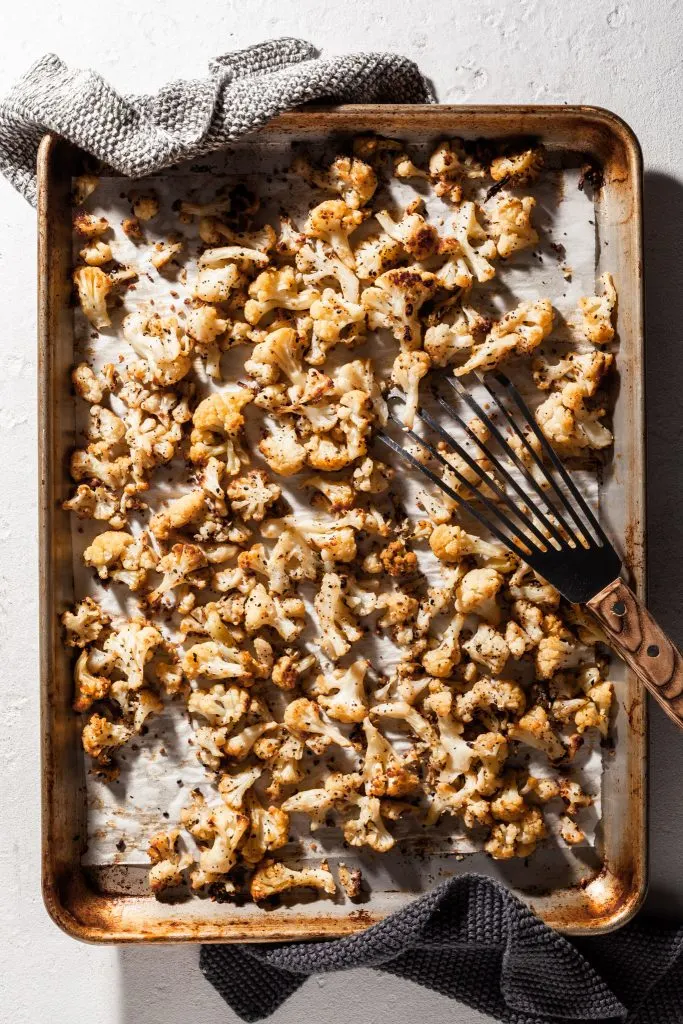 An aluminum baking sheet lined with parchment paper is covered with roasted cauliflower coated in seasonings; a wood handled spatula rests on the pan and knit pot holders have been left on the sides of the sheet. 