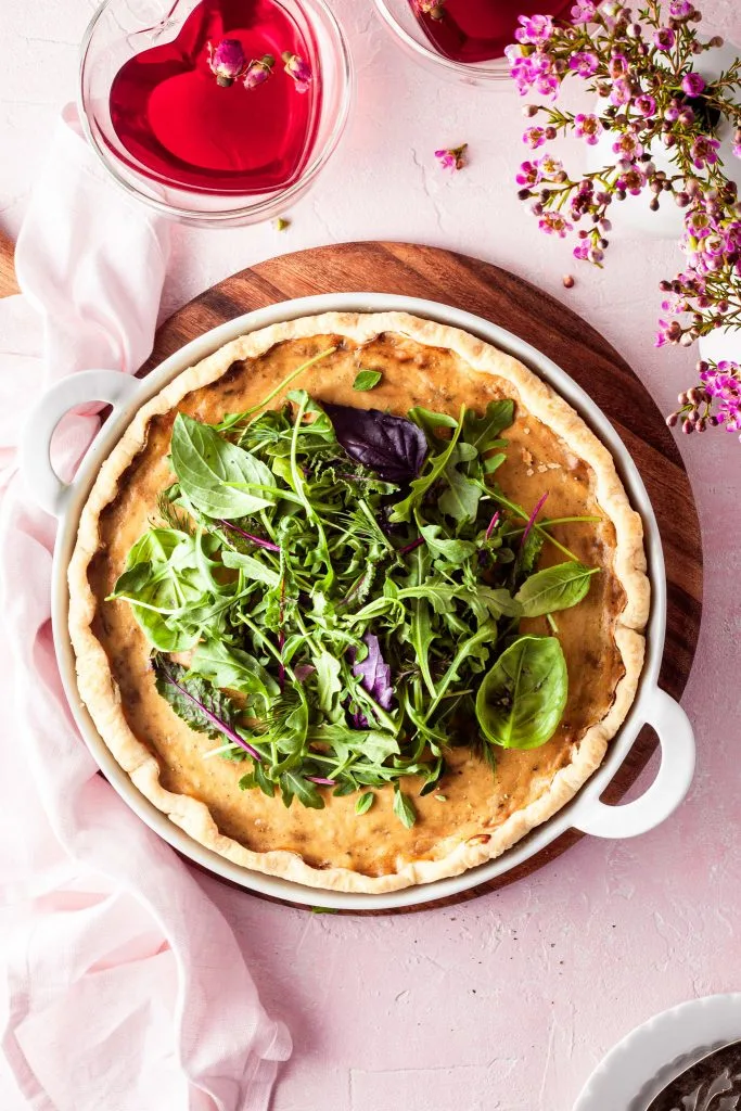 Flatlay of a quiche top in a white baking dish resting on a  wooden board, beside a mug of rose tea. 