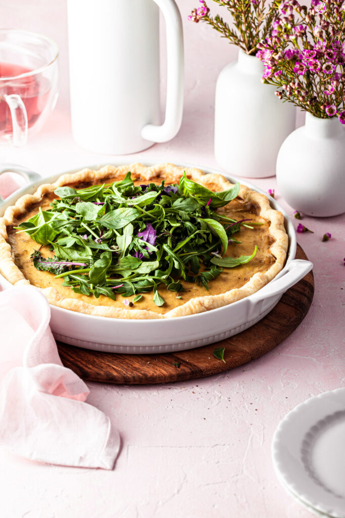 Side view of a vegan french onion quiche in a white porcelain baking dish. In the background there is a white French press and a mug of tea.
