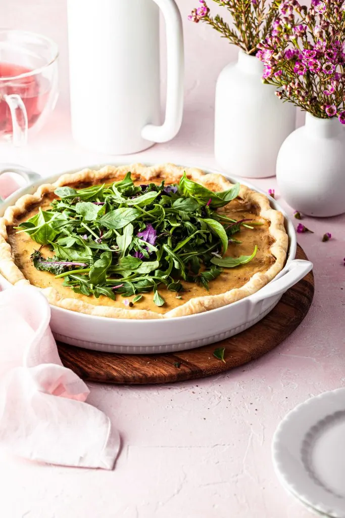 Side view of a vegan french onion quiche in a white porcelain baking dish. In the background there is a white French press and a mug of tea.