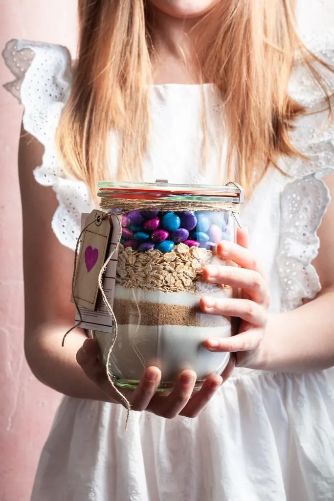 A little girl with long blond hair handing a gift of cookie mix in a glass jar. 