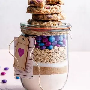 A jar filled with cookie mix, topped with baked vegan m&m cookies.