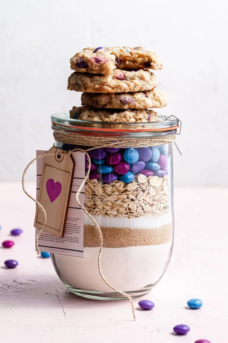 A jar filled with cookie mix, topped with baked vegan m&m cookies.