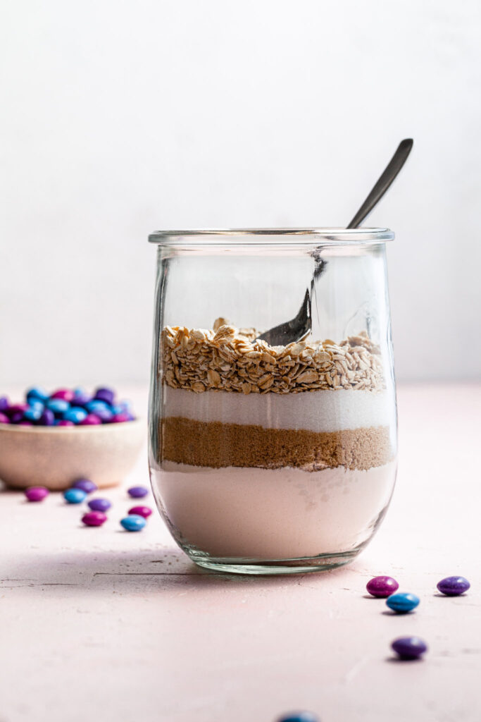 A side view of a canning jar partly filled with flour, sugar and oats for cookie mix. In the background there is a small bowl of candies.