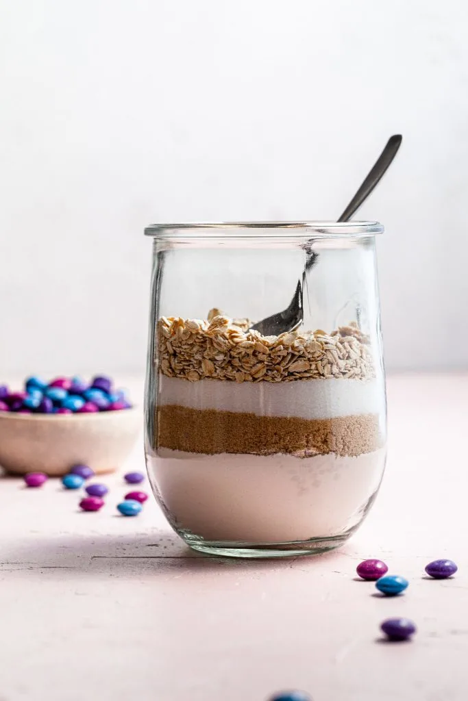 A side view of a canning jar partly filled with flour, sugar and oats for cookie mix. In the background there is a small bowl of candies.