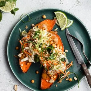 View looking down on a green plate filled with a sweet potato cut in half, topped with cripsy chickpeas, peanut sauce and coleslaw.