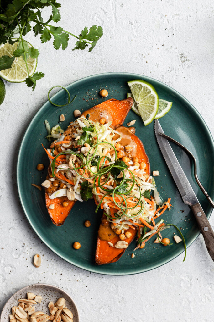 View looking down on a green plate filled with a sweet potato cut in half, topped with cripsy chickpeas, peanut sauce and coleslaw.
