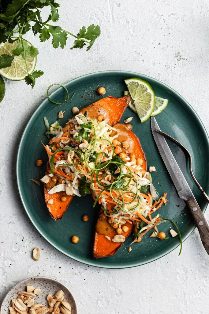 View looking down on a green plate filled with a sweet potato cut in half, topped with cripsy chickpeas, peanut sauce and coleslaw.
