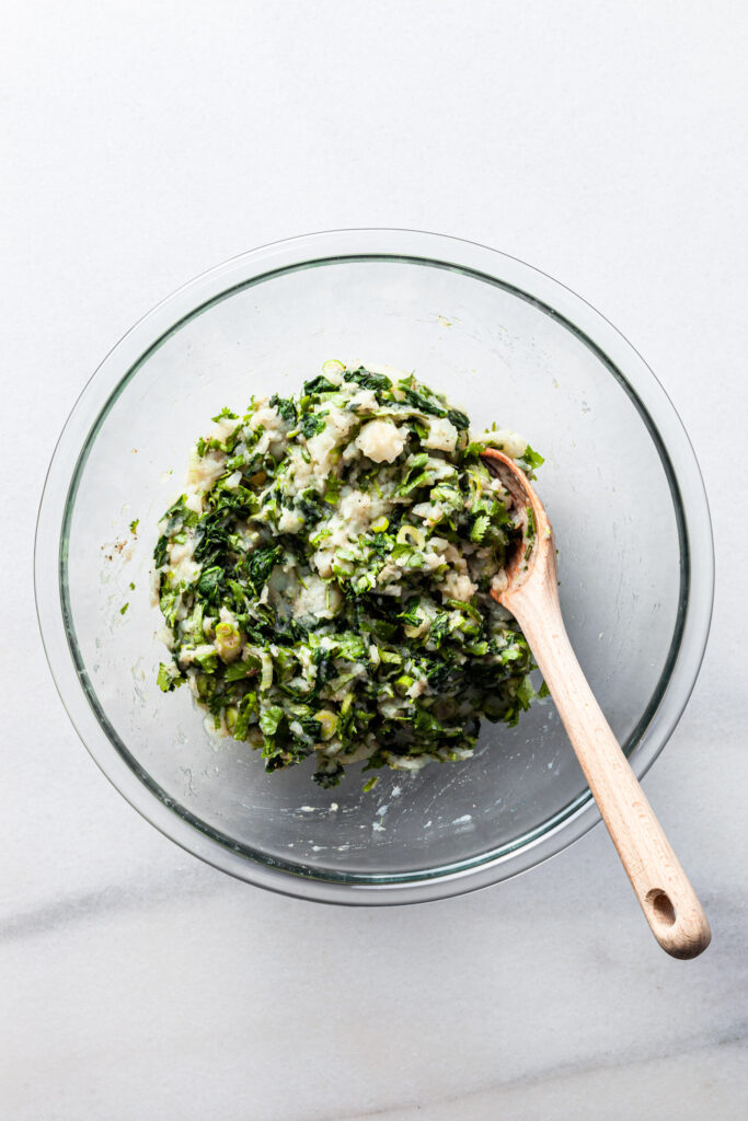 A glass bowl filled with a mixture of mashed potatoes, chopped spinach, sliced green onions, cilantro and spices. 