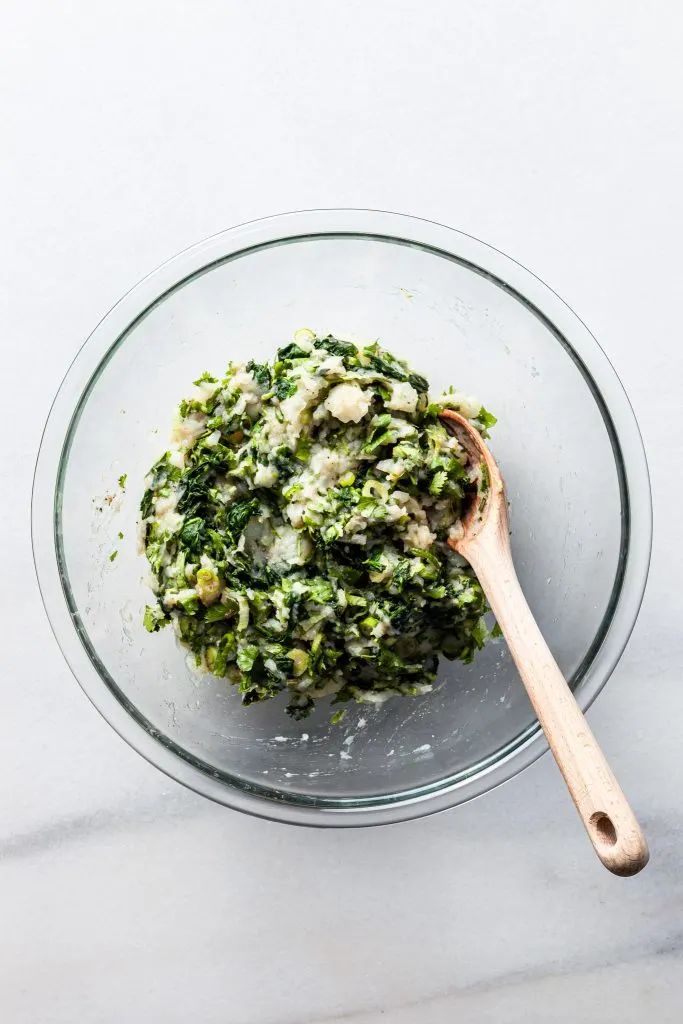 A glass bowl filled with a mixture of mashed potatoes, chopped spinach, sliced green onions, cilantro and spices. 