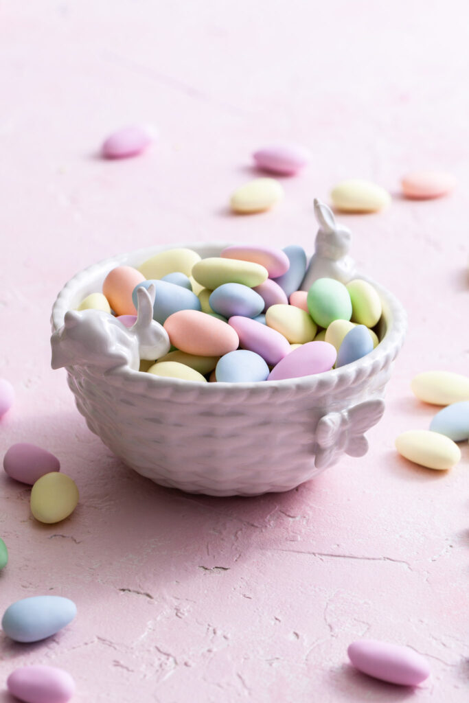 A small white bowl decorated with small rabbits and a butterfly is filled with pastel Easter candies
