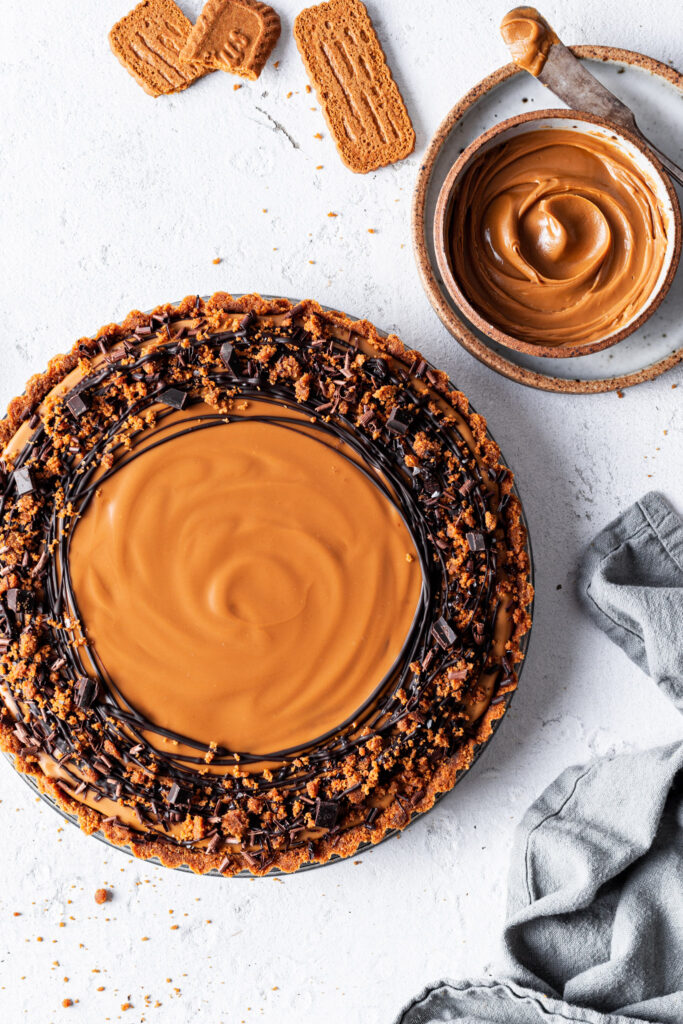 A chocolate speculoos tart not yet sliced, garnished with drizzled chocolate beside a small bowl of cookie butter and Biscoff cookies. 
