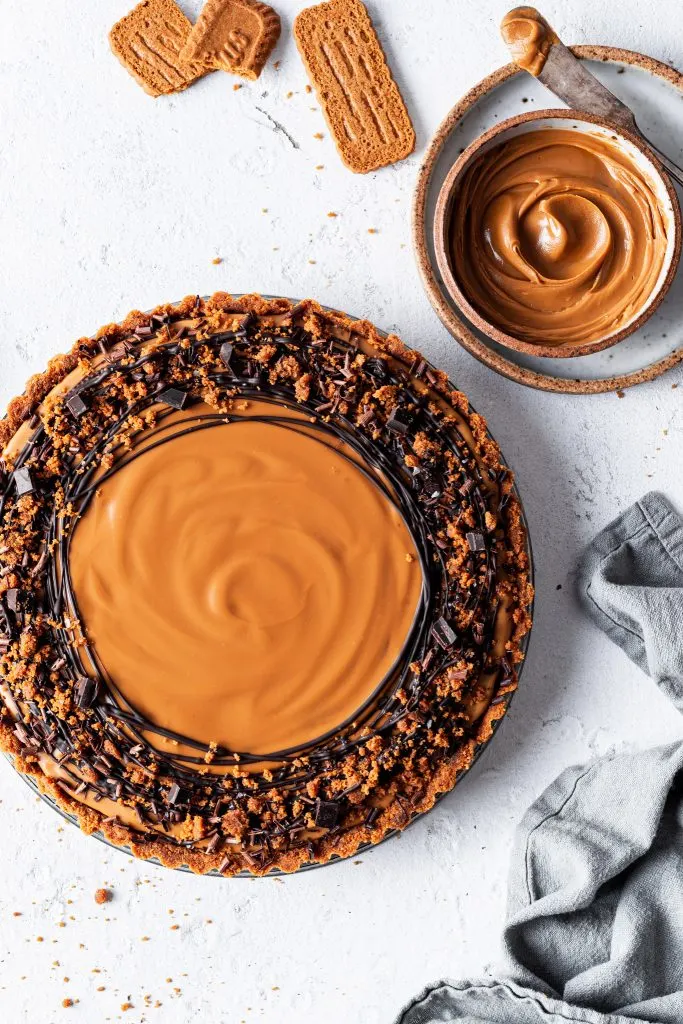 A chocolate speculoos tart not yet sliced, garnished with drizzled chocolate beside a small bowl of cookie butter and Biscoff cookies. 
