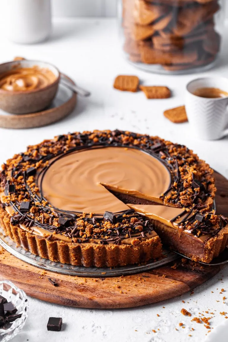 A Vegan chocolate biscoff tart with one slice cut and partly removed to reveal the inside of the tart showing off the chocolate layer topped with cookie butter.