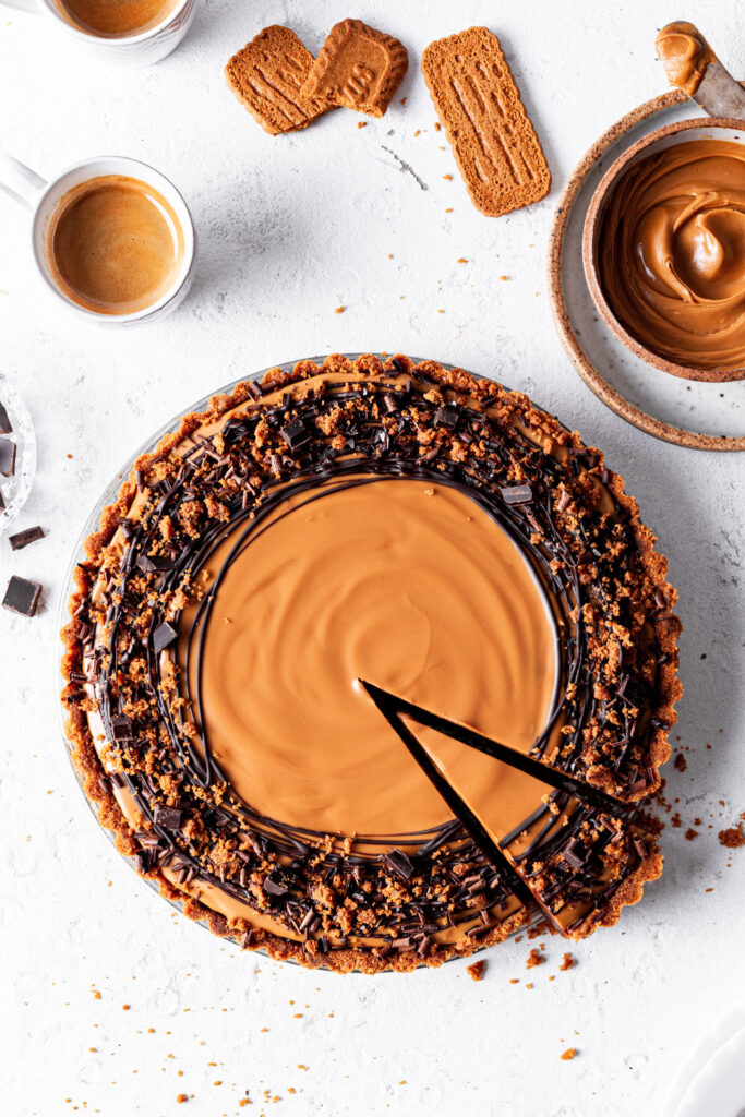 A chocolate cookie tart with one clean slice cut, beside a cup of espresso, a small bowl of cookie butter and some speculoos cookies. 