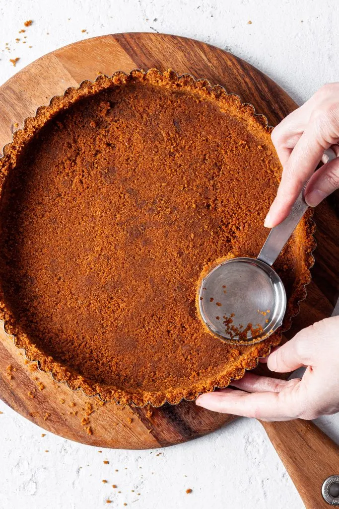 Hands using a measuring cup to press a cookie crumb crust into a tart pan.