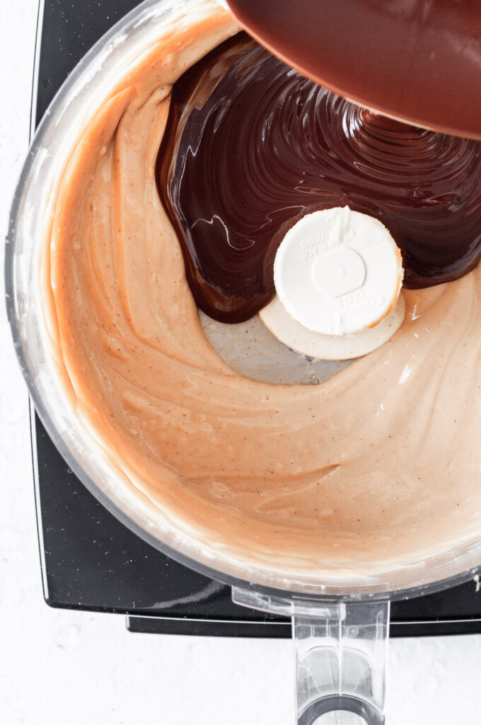 View into a food processor where melted chocolate is being added to the tofu and cookie butter mixture. 