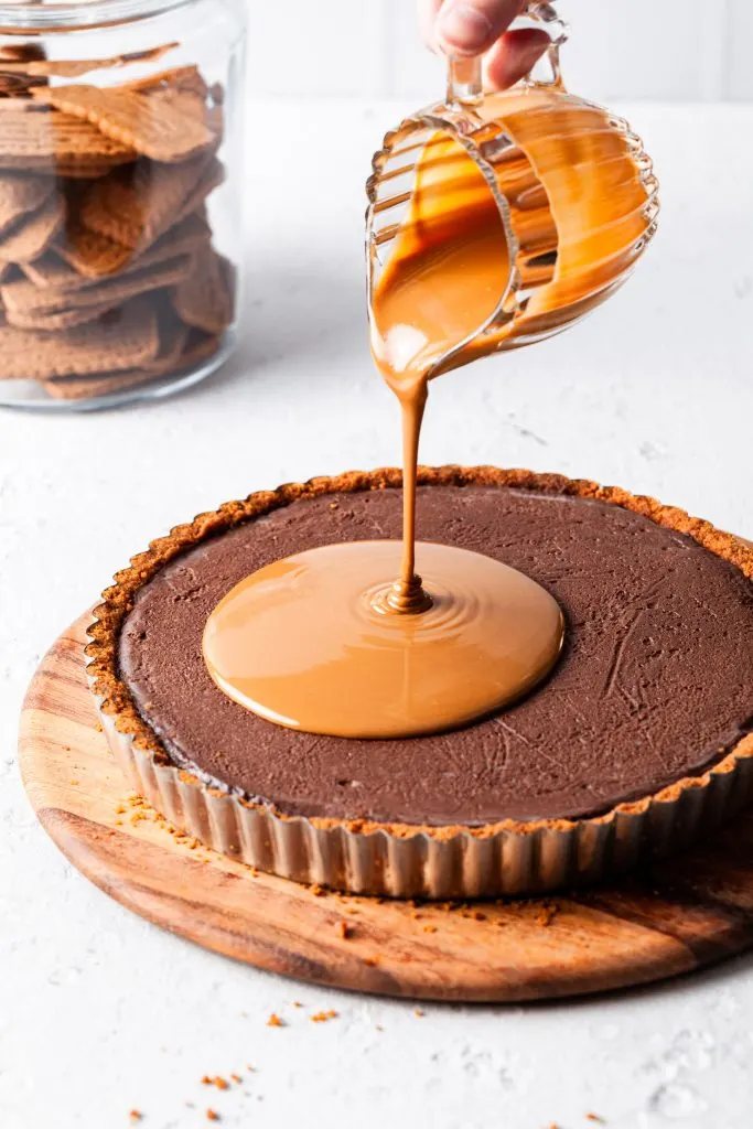 A small glass jug filled with melted Biscoff spread is pouring over the chocolate cookie tart as the final layer. 