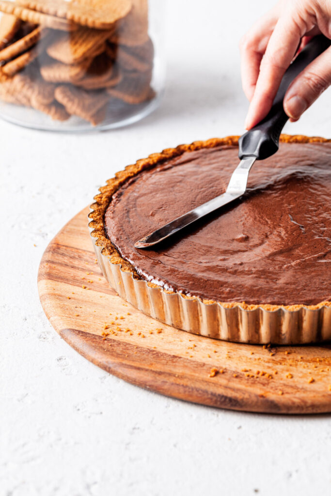 An image of a hand holding an offset spatula smoothing the chocolate Biscoff filling over the cookie crumb crust before chilling. 