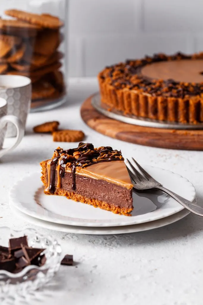 A slice of chocolate cookie butter tart on a white plate, drizzled with melted dark chocolate and cookie butter. 