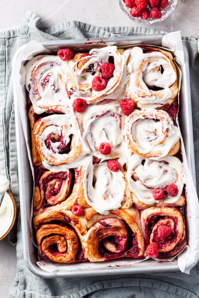 A baking tray filled with raspberry scrolls after baking, 8 out of 12 of them have been frosted with vanilla glaze. 