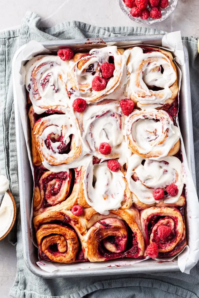 A baking tray filled with raspberry scrolls after baking, 8 out of 12 of them have been frosted with vanilla glaze. 