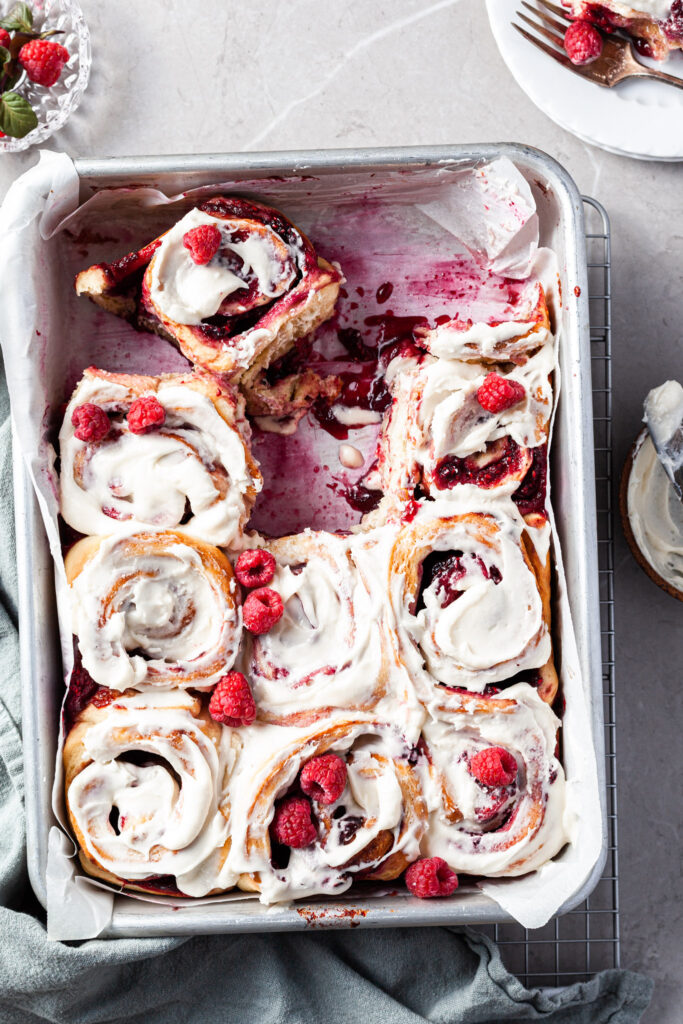 Raspberry sweet buns in a baking tray, 3 are missing and a small plate sits at the corner with a roll on it.