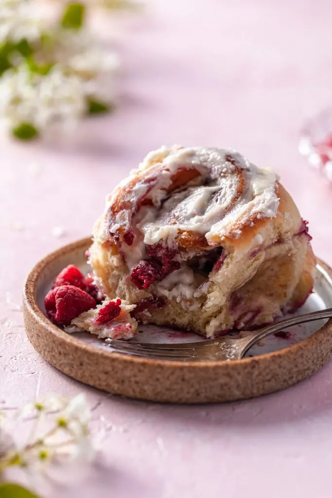 A single raspberry sweet bun with a bite missing sits on an artisan ceramic plate. White blossoms are bokahed in the background. 