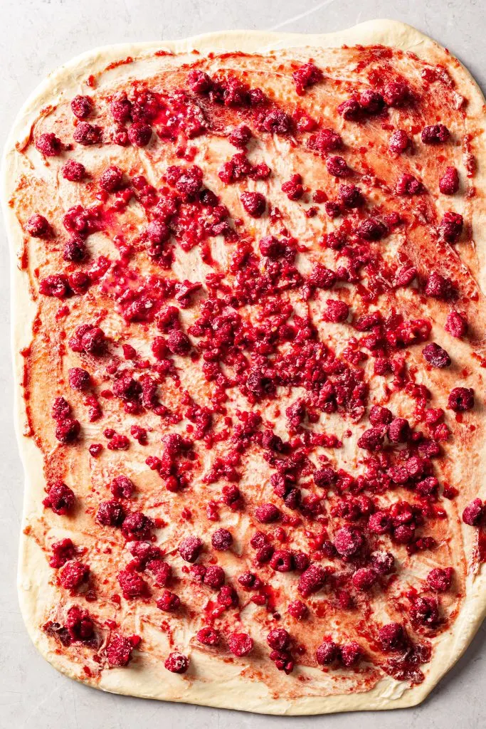 Sweet dough rolled out with vegan butter, raspberry preserves and frozen raspberries spread out on the top. 