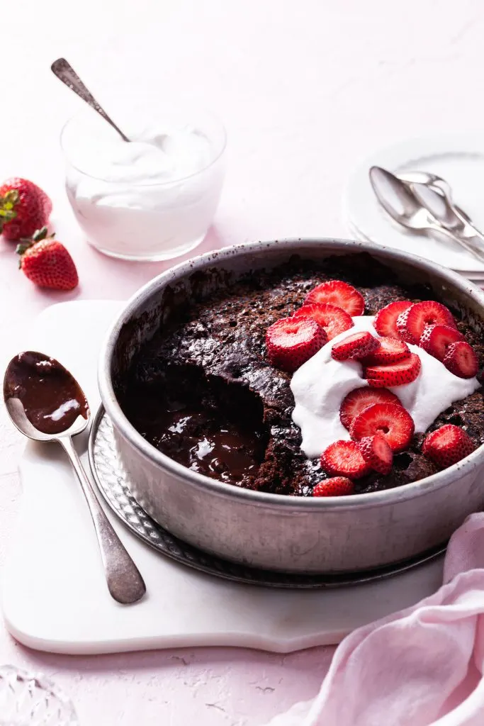 A round cake pan filled with a partially served chocolate cake exposing chocolate sauce on the bottom. Garnished with whipped cream and sliced strawberries.
