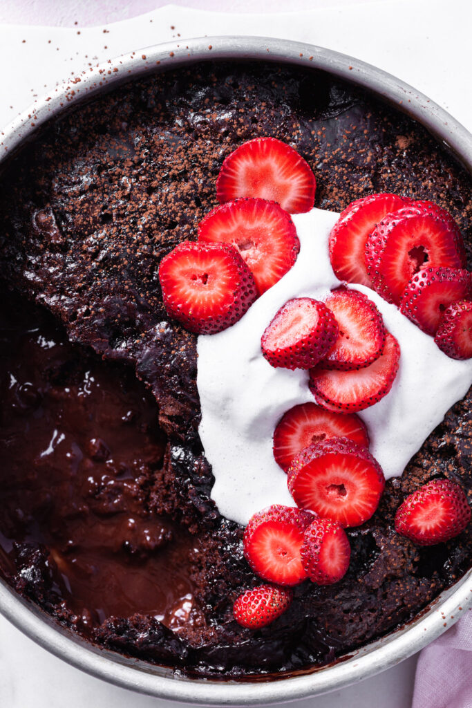 Close up of the self-saucing chocolate strawberry pudding cake with a serving missing so that both the cake and the sauce are visible. The top of the vegan dessert is topped with non-dairy whip and fresh sliced strawberries. 
