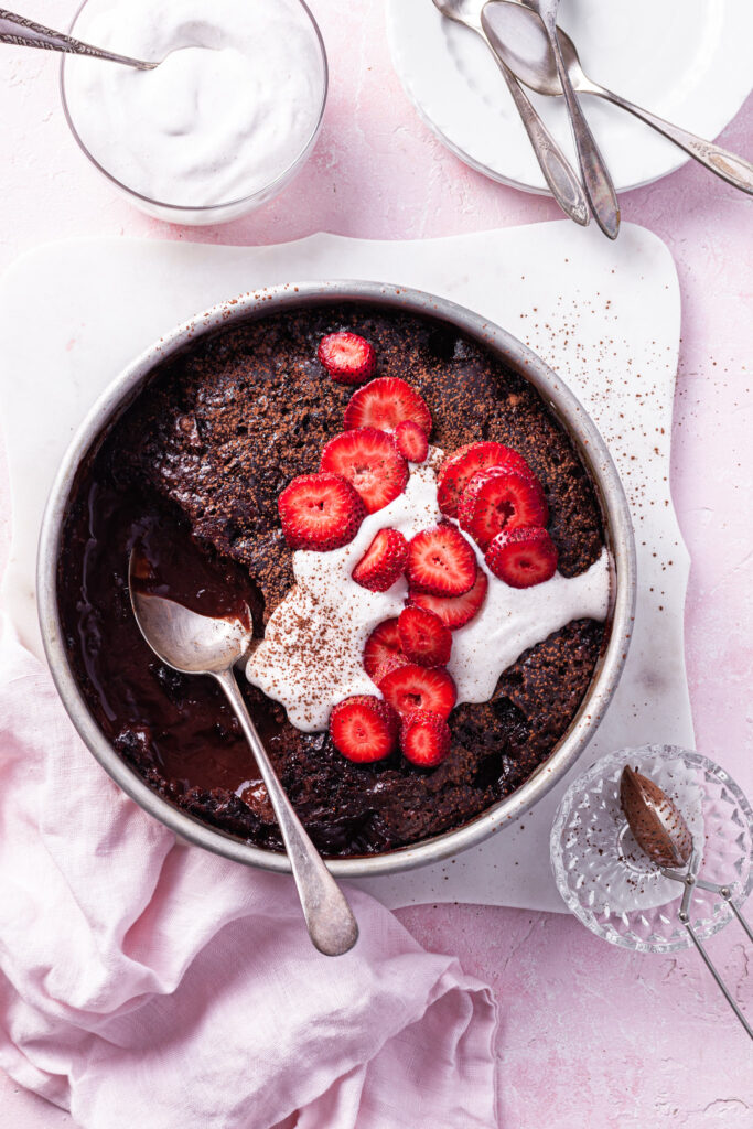 A pink counter with the self-saucing chocolate strawberry pudding partly served beside a bowl of vegan whipped cream.