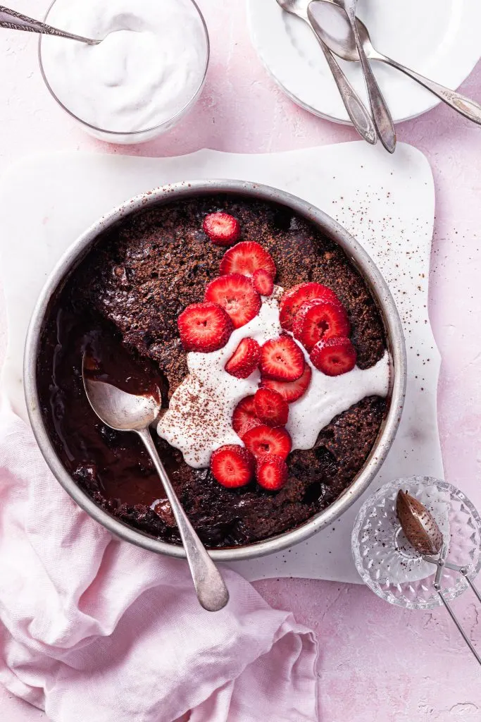 A pink counter with the self-saucing chocolate strawberry pudding partly served beside a bowl of vegan whipped cream.