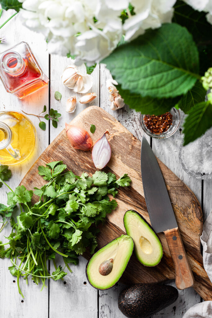 An image of the ingredients for the avocado chimichurri sauce on a table in summer sunlight with a cutting board and kitchen knife.