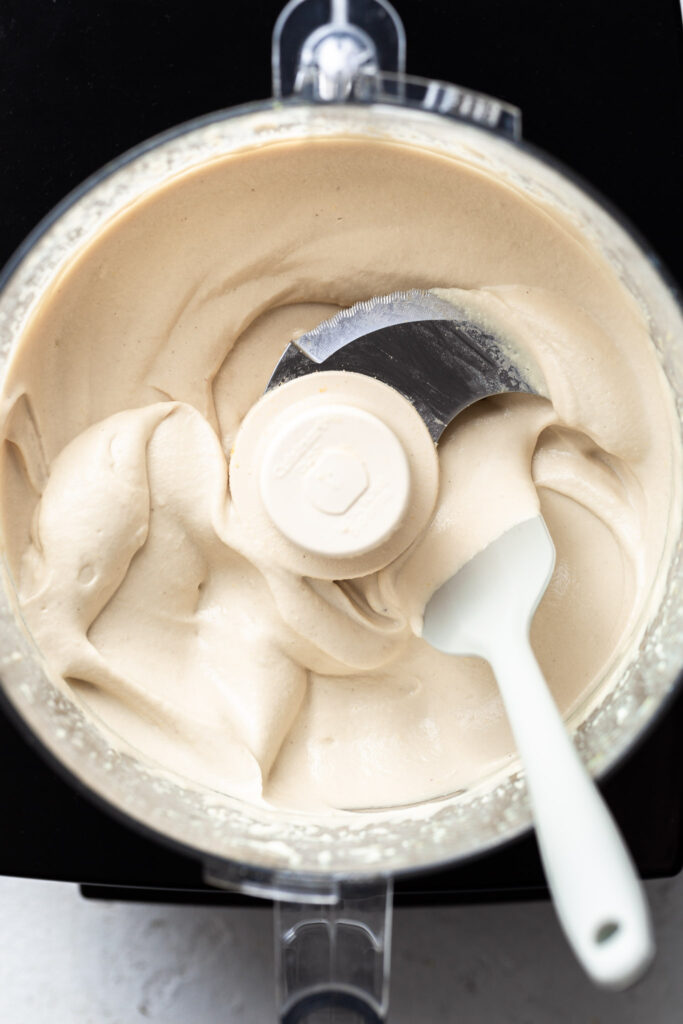 A food processor filled with off-white cashew ricotta which appears to be satiny in texture. 