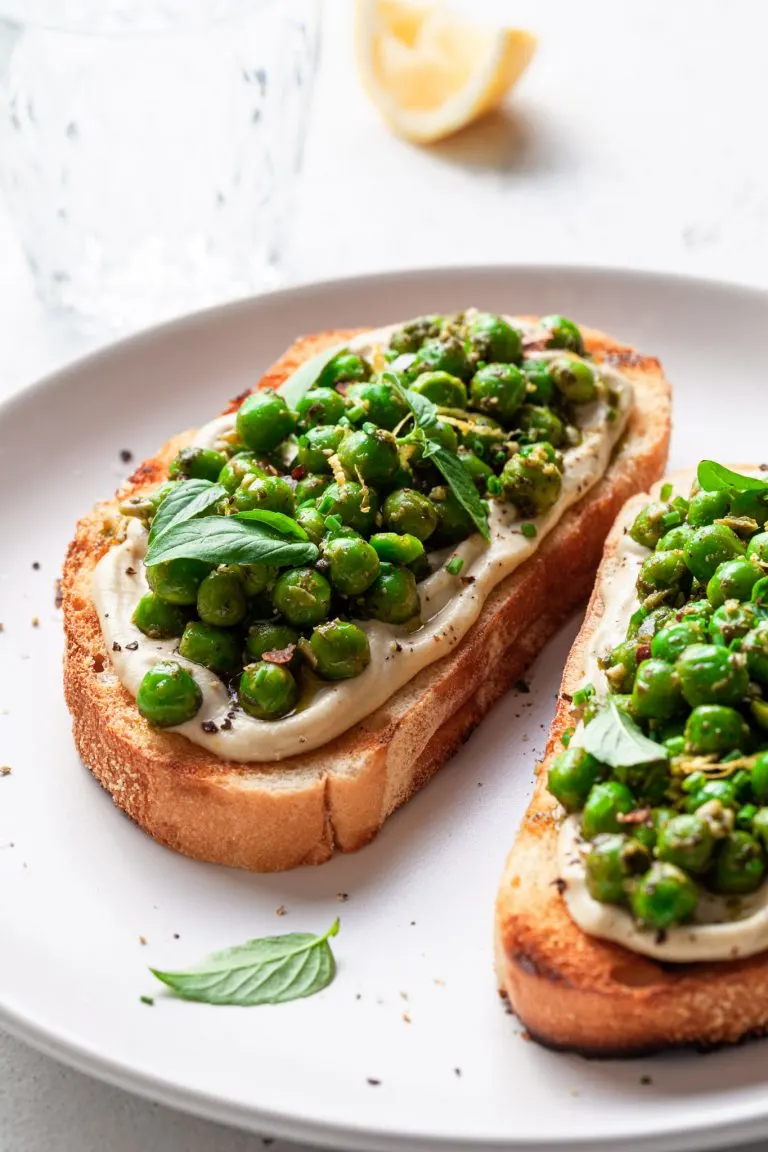 Two slices of toast on a white plate, topped with whipped vegan ricotta and green peas covered in pesto. A lemon wedge is in the background beside a glass of water.