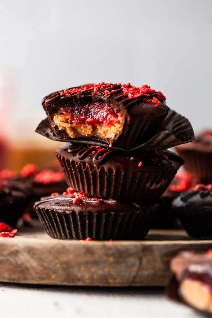 A stack of peanut butter jelly cups on a wooden plate, the top cup has a bite taken out of it revealing the layered filling. 