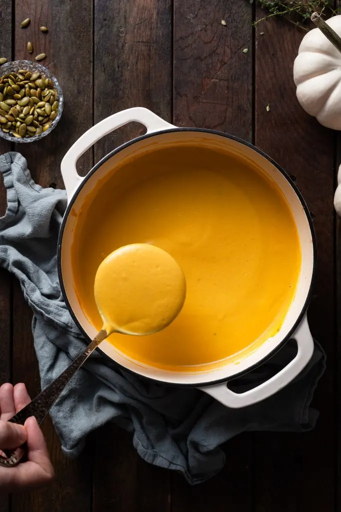 A white Dutch oven on a dark wooden table filled with creamy pumpkin soup. 