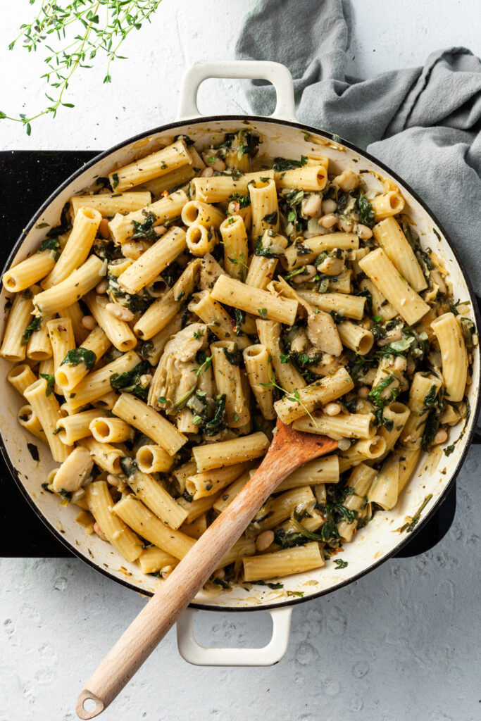 A white enamel skillet filled with noodles, cooked spinach, white beans and artichokes. 