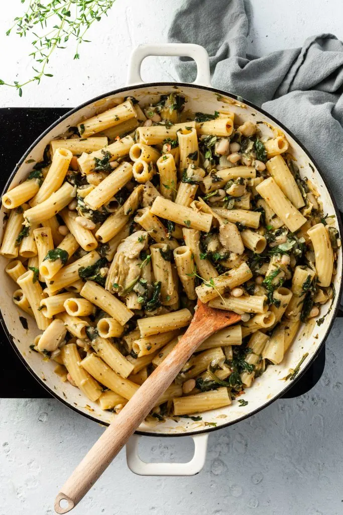 A white enamel skillet filled with noodles, cooked spinach, white beans and artichokes. 