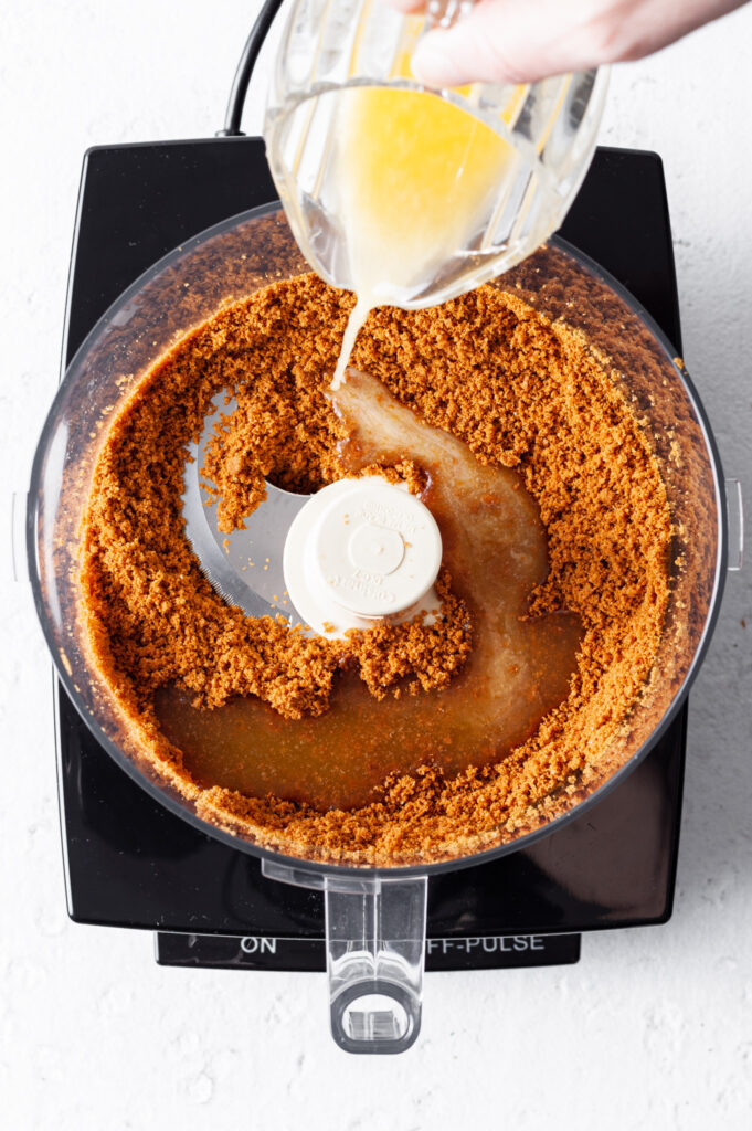 Food processor with cookie crumbs in base and melted vegan butter being drizzled in