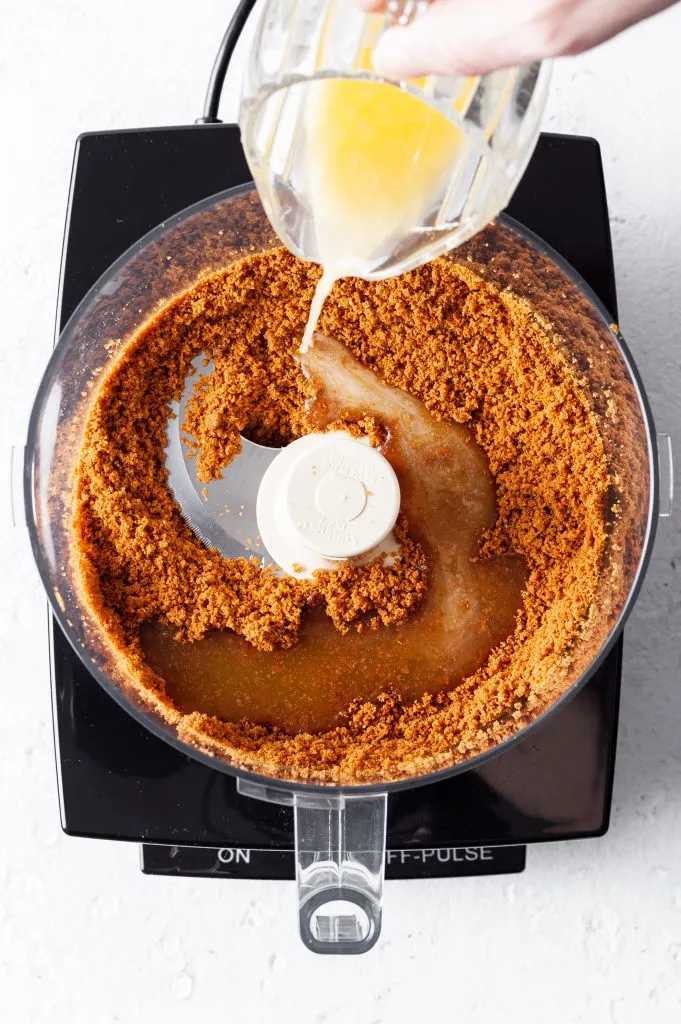 Food processor with cookie crumbs in base and melted vegan butter being drizzled in