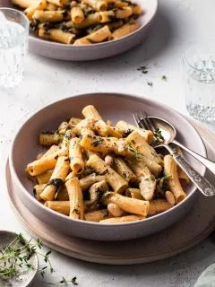 A shallow bowl is filled with medium tubes of pasta, speckled with cooked spinach, white beans, and artichokes.