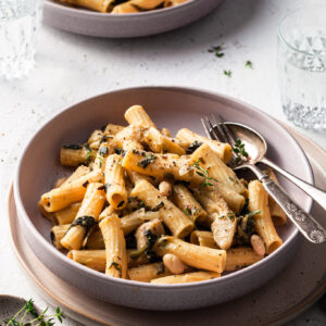 A shallow bowl is filled with medium tubes of pasta, speckled with cooked spinach, white beans, and artichokes.