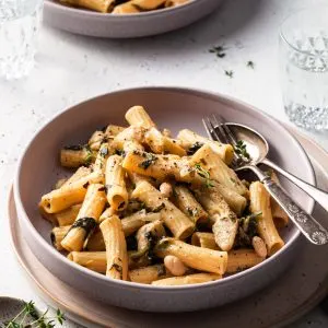 A shallow bowl is filled with medium tubes of pasta, speckled with cooked spinach, white beans, and artichokes.