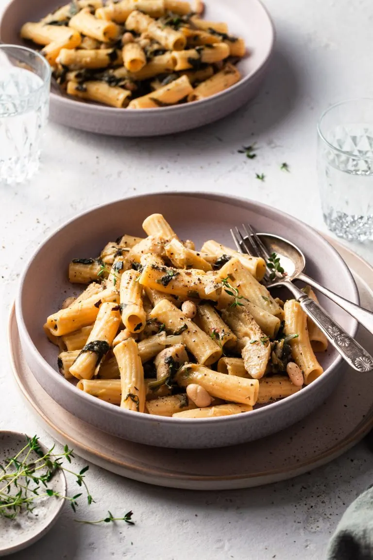 A shallow bowl is filled with medium tubes of pasta, speckled with cooked spinach, white beans, and artichokes.